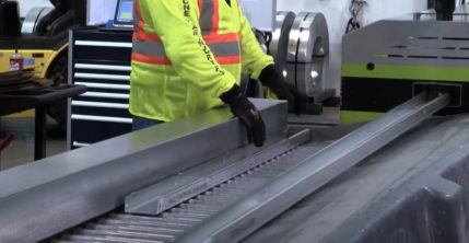 Employee wearing safety vest and gloves reaching for piece of metal on rollers