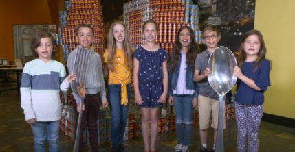 Group of seven kids holding large spoon and fork in front of large stacks of canned food